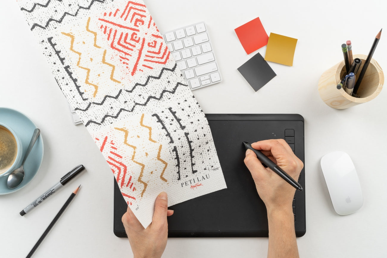 A pair of hands is shown using a tablet and stylus pen. One hand holds wax block print like artwork by artist Peti Lau. There are red, black, and yellow paint chips on the desk.