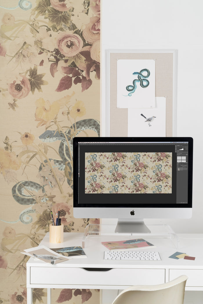 A computer showing a repeat pattern of a snake and flowers in Adobe Photoshop. The original snake motif painting by artist Ashley Woodson Bailey is hanging on the wall in the background. The final printed wallpaper is also on the wall.