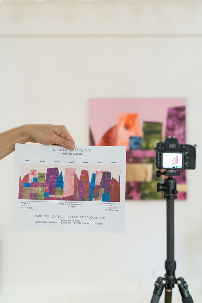 In the foreground a hand holds a panel map showing a complete mural. In the background the original artwork the mural was made from is being photographed. The mural is large colorful blocks that mimic a city skyline, by artist Alexandra Valenti.